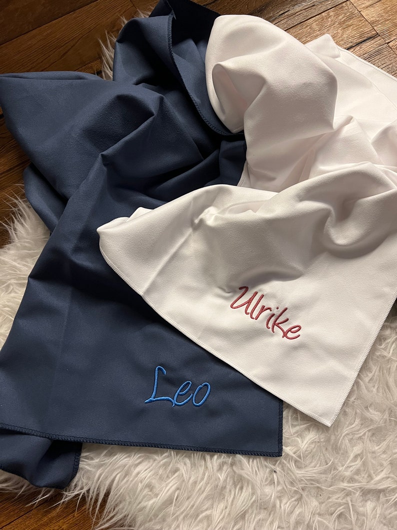 May include: Two personalized towels, one navy blue with the name "Leo" embroidered in blue thread, and one white with the name "Ulrike" embroidered in red thread.