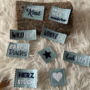 Könnte beinhalten: Ein Set aus neun blauen und weißen gestickten Bügeletiketten mit verschiedenen Sprüchen, darunter "Wunsch Kind", "Frech Wild Wunderbar", "Wildfang", "Frech Dachs", "Wunderbar", "Cool Kids", "Herz Stück", ein Stern und ein Herz.