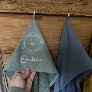 May include: Three hooded towels hanging on hooks. The towel on the left is white, the middle towel is green with a white embroidered lion and the name "Emilian", and the towel on the right is blue.