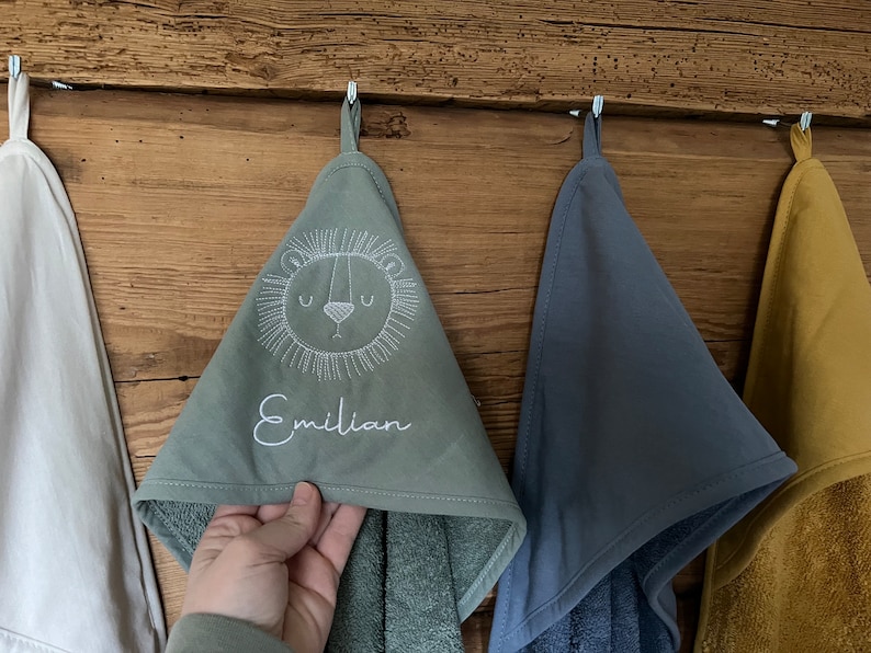May include: Three hooded towels hanging on hooks. The towel on the left is white, the middle towel is green with a white embroidered lion and the name "Emilian", and the towel on the right is blue.