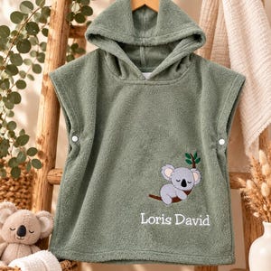 Può includere: Un poncho da bagno con cappuccio color verde salvia in morbida spugna. La parte anteriore presenta un ricamo di un koala che dorme su un ramo e la scritta bianca Loris David. I lati sono dotati di bottoni a pressione bianchi per la chiusura.