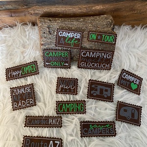 Può includere: Una collezione di toppe in feltro marrone con cuciture bianche, che presentano testo e grafica a tema campeggio. Le toppe includono frasi come "Happy Camper", "Camping Makes You Happy", "Save Campers", e "Camper Parking Only".