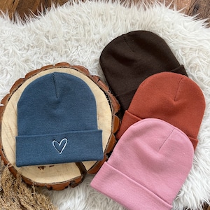 Könnte beinhalten: Vier Strickmützen in verschiedenen Farben: Braun, Blau, Orange und Rosa. Die blaue Mütze hat ein weißes Herz auf der Vorderseite gestickt.