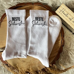 Könnte beinhalten: Weiße gerippte Socken mit dem Text "BESTE Schwester" in schwarzer Schrift. Die Socken werden auf einer Holzscheibe mit flauschigem weißem Hintergrund präsentiert. Ein kleines Holzschild ist sichtbar.