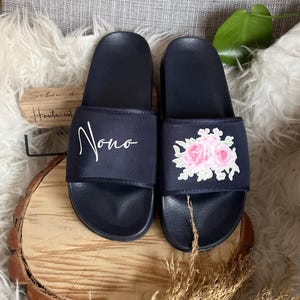 May include: A pair of navy blue slides with a floral embroidery design on one slide and the word "Nono" embroidered on the other slide.
