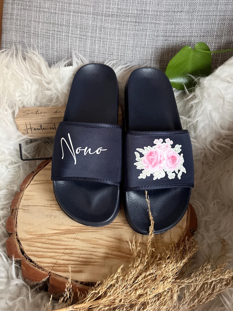 May include: A pair of navy blue slides with a floral embroidery design on one slide and the word "Nono" embroidered on the other slide.