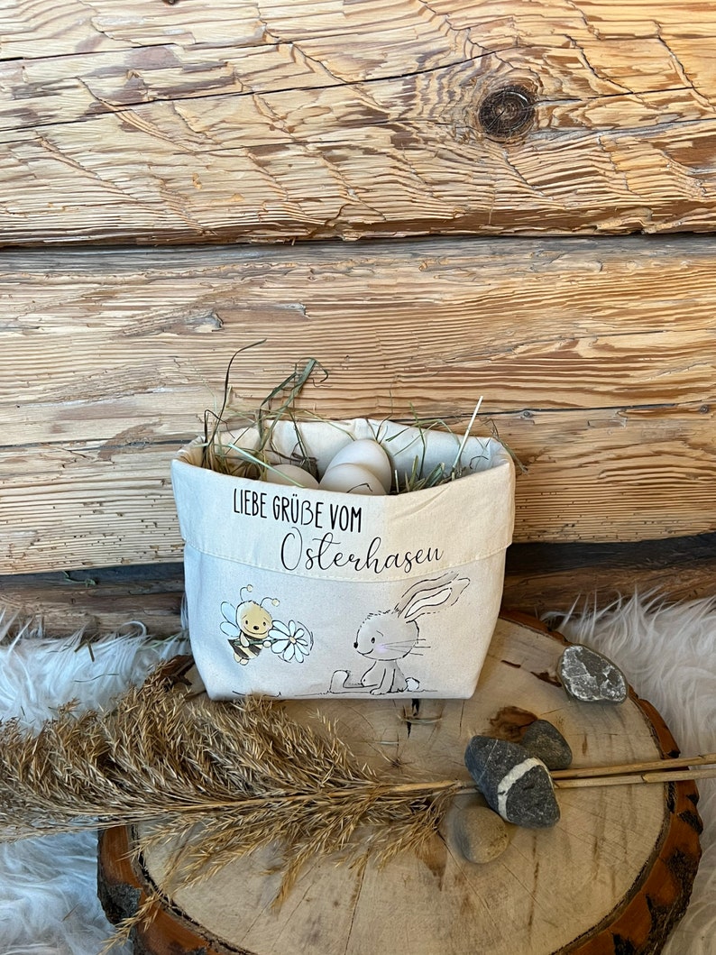 K&ouml;nnte beinhalten: Ein wei&szlig;er Stoff-Osterkorb mit einem Hasen- und Bienen-Design. Der Korb ist mit Stroh und wei&szlig;en Eiern gef&uuml;llt. Der Text auf dem Korb lautet "Liebe Gr&uuml;&szlig;e vom Osterhasen".