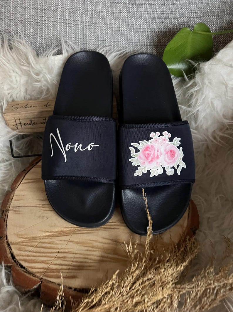 May include: A pair of black slides with a floral design on one slide and the word "Nono" on the other slide.