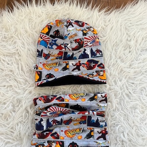 Puede incluir: Un gorro y un cuello de lana gris y negro con un estampado de cómic con superhéroes y las palabras "POW!!!", "BOOM!" y "SPLASSH!"