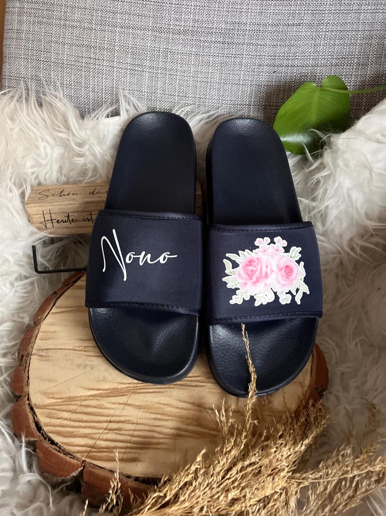 May include: Pair of black slide sandals with embroidered floral designs. One sandal has the word "Nono" embroidered on it.