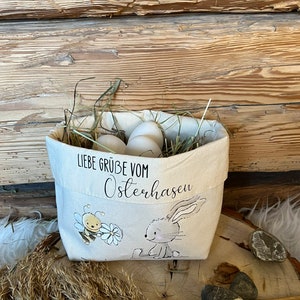K&ouml;nnte beinhalten: Eine wei&szlig;e Stofftasche mit einem Hasen- und Bienenmotiv. Die Tasche ist mit Stroh und wei&szlig;en Eiern gef&uuml;llt. Auf der Tasche steht "Liebe Gr&uuml;&szlig;e vom Osterhasen".