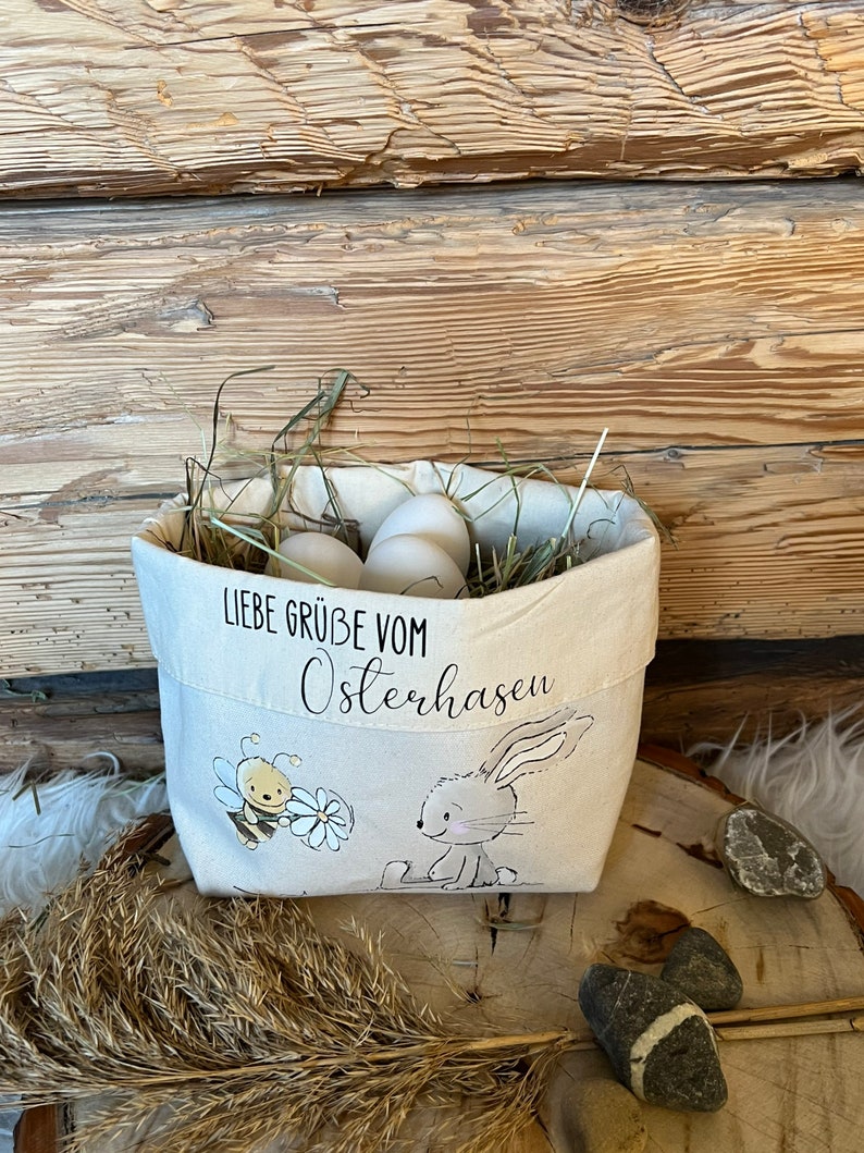 K&ouml;nnte beinhalten: Eine wei&szlig;e Stofftasche mit einem Hasen- und Bienenmotiv. Die Tasche ist mit Stroh und wei&szlig;en Eiern gef&uuml;llt. Auf der Tasche steht "Liebe Gr&uuml;&szlig;e vom Osterhasen".