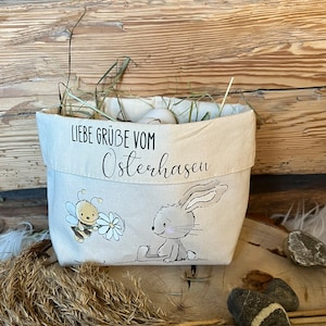 K&ouml;nnte beinhalten: Ein wei&szlig;er Canvas-Osterkorb mit einer Hasen- und Bienen-Illustration. Der Korb ist mit Stroh gef&uuml;llt und hat den Text "Liebe Gr&uuml;&szlig;e vom Osterhasen" auf der Vorderseite.