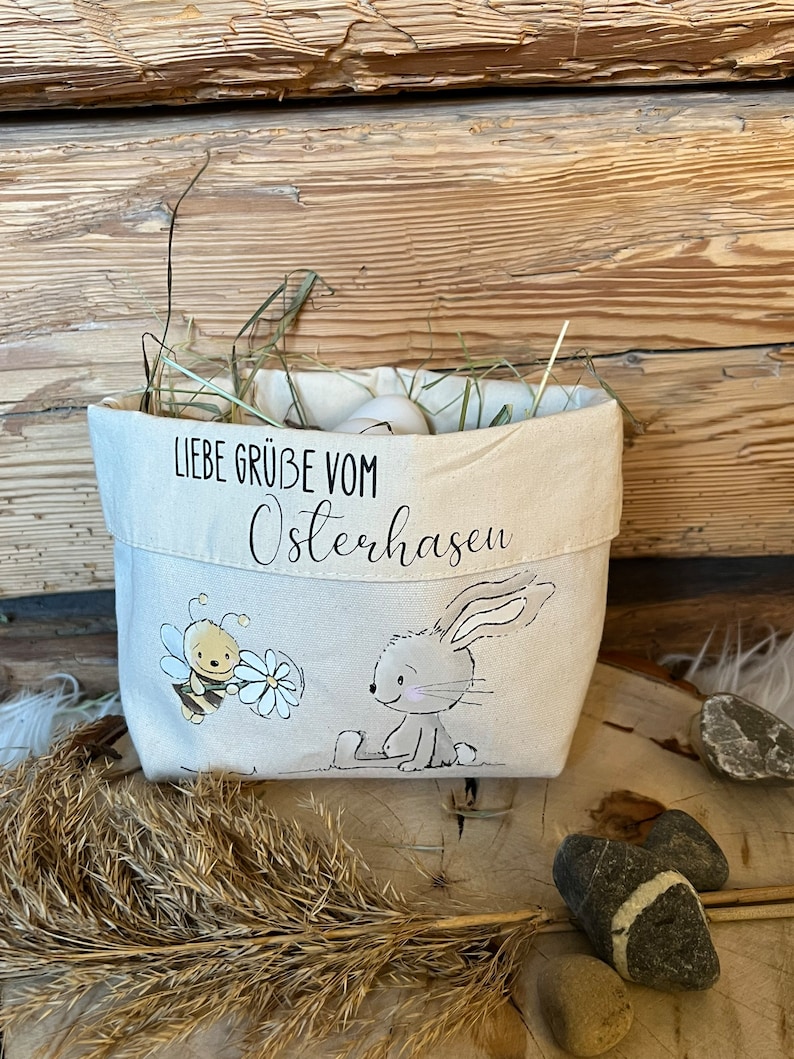 K&ouml;nnte beinhalten: Ein wei&szlig;er Canvas-Osterkorb mit einer Hasen- und Bienen-Illustration. Der Korb ist mit Stroh gef&uuml;llt und hat den Text "Liebe Gr&uuml;&szlig;e vom Osterhasen" auf der Vorderseite.