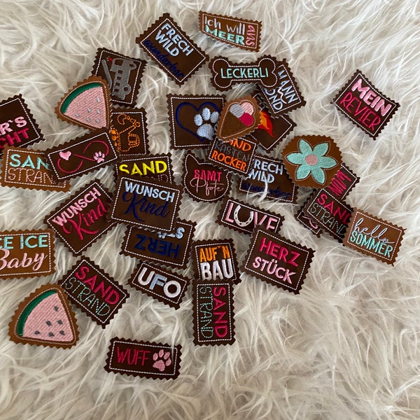 May include: A collection of brown leather patches with various embroidered designs and text in German. The patches feature words like "Sand", "Wunsch", "Frech", "Wild", "Love", "Herz", "UFO", "Sommer", and "Mein Revier".