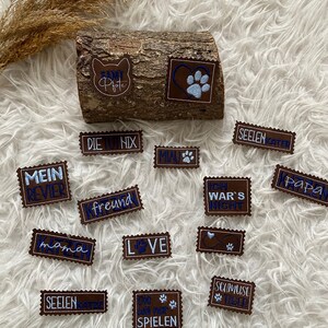 Könnte beinhalten: Eine Sammlung von braunen Leder-Patches mit weißen und blauen Stickereien. Die Patches zeigen verschiedene Sätze auf Deutsch, darunter "Mein Revier", "Die Nix", "K-Freund", "Love", "Seelenkatze", "Ich War's Nicht", "Schmuse Tiger", "Papa" und "Die Will Nur Spielen".