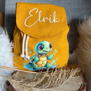 Könnte beinhalten: Ein gelber Canvas-Rucksack in Senfgelb mit einem weißen Kordelzug und einem niedlichen Comic-Schildkröten-Motiv. Der Rucksack trägt den Namen "Elrik" in weißer Kursivschrift auf der Vorderseite.