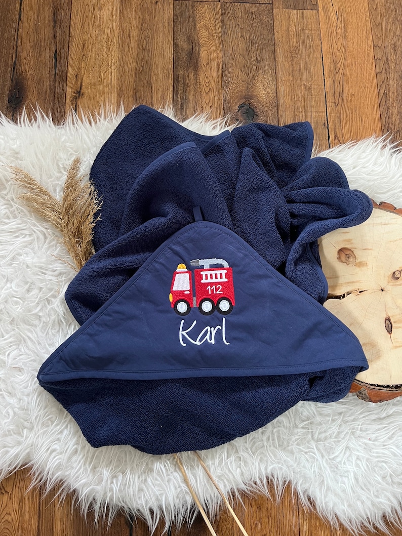 May include: A navy blue hooded towel with a red fire truck embroidered on the front. The fire truck has the number 112 on the side and the name "Karl" embroidered below it.