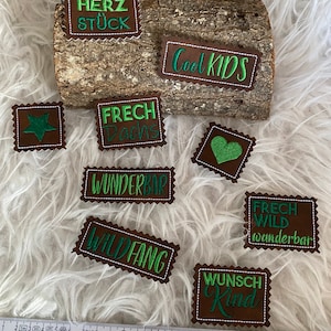 Könnte beinhalten: Neun braune Filzpatches mit grünem Sticktext. Die Patches beinhalten die Wörter "Herz Stück", "Cool Kids", "Frech Dachs", "Wunderbar", "Wild Fang", "Frech Wild wunderbar", "Wunsch Kind" und zwei Patches mit einem Stern und einem Herzen.