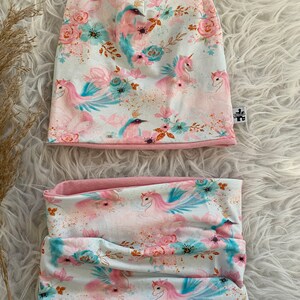 Könnte beinhalten: Ein pink- und blaues Mützen- und Halssocken-Set mit Einhorn-Druck. Der Stoff hat einen weißen Hintergrund mit pinkfarbenen und blauen Einhörnern, Blumen und goldenem Glitzer.
