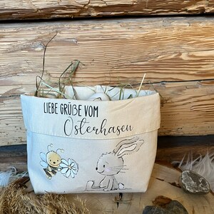 K&ouml;nnte beinhalten: Eine wei&szlig;e Stofftasche mit einer Hasen- und Bienen-Illustration. Die Tasche hat den Text "Liebe Gr&uuml;&szlig;e vom Osterhasen" aufgedruckt.
