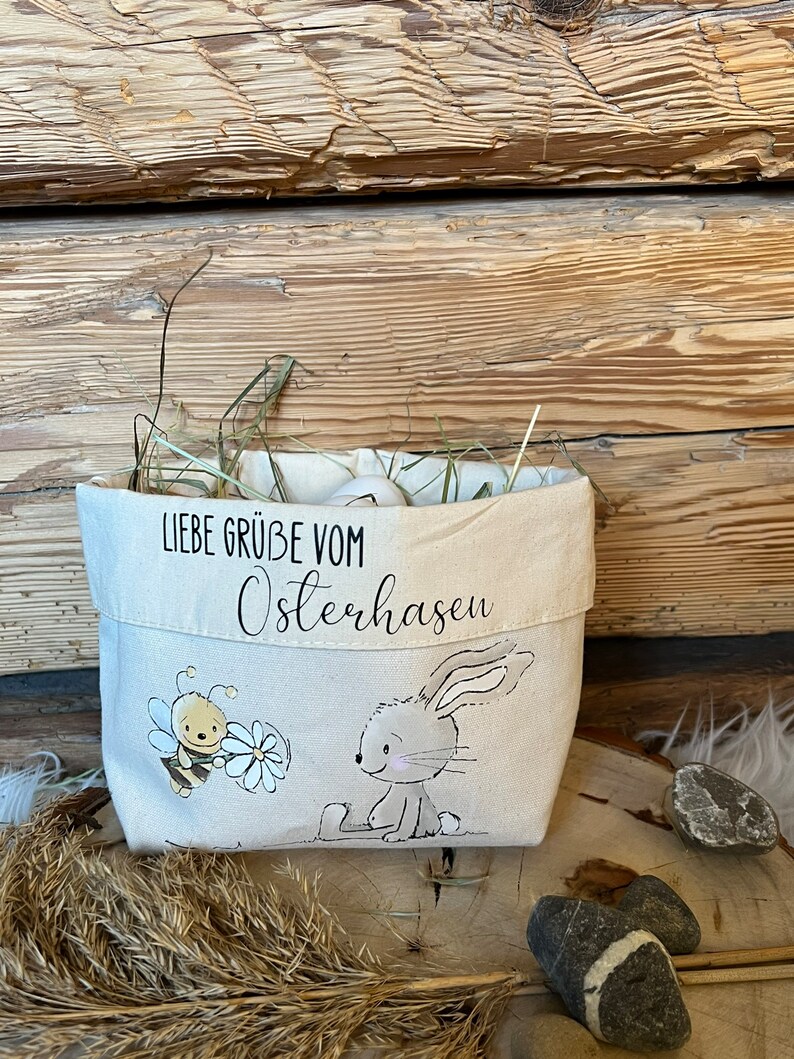 K&ouml;nnte beinhalten: Eine wei&szlig;e Stofftasche mit einer Hasen- und Bienen-Illustration. Die Tasche hat den Text "Liebe Gr&uuml;&szlig;e vom Osterhasen" aufgedruckt.