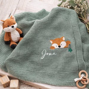 Könnte beinhalten: Eine salbeigrüne Strickdecke mit einem gestickten Fuchs, der über einer Tasche hervorlugt, und dem Namen Jona in weißer Schreibschrift. Ein kleiner Plüschfuchs, zwei hölzerne Buchstabenwürfel und ein hölzerner Greifring liegen bei der Decke.