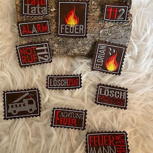 Può includere: Una collezione di toppe in feltro marrone con ricami bianchi e rossi. Le toppe presentano vari disegni a tema pompieri, tra cui le parole "FEUER", "ALARM", "NOT RUF", "LÖSCHZUG", "ACHTUNG FEUER", "LOSCH MEISTER", "FLORIAN", "FEUER WEHR MANN", "TATÜ TATA" e "112".