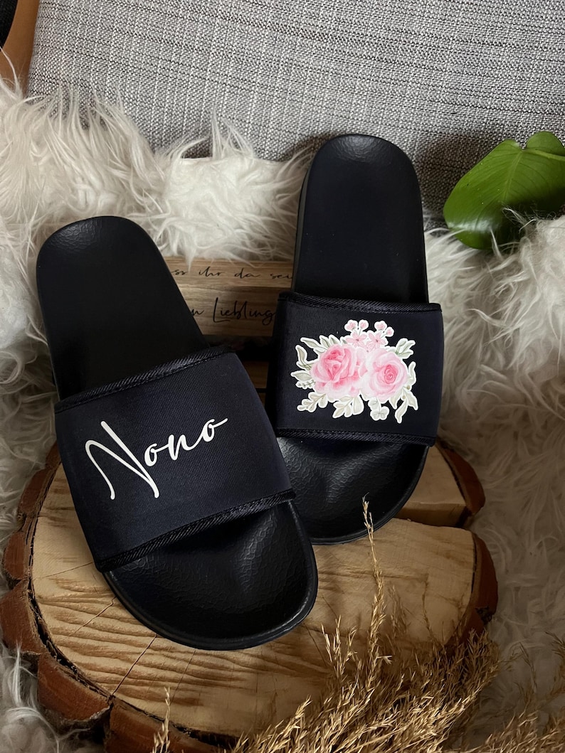 May include: A pair of black slides with a white floral design on one slide and the word "Nono" on the other slide.