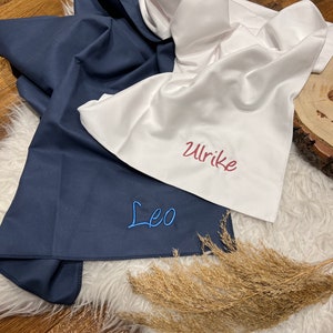 May include: Two personalized fabric squares, one navy blue with the name "Leo" embroidered in blue thread, and one white with the name "Ulrike" embroidered in red thread.
