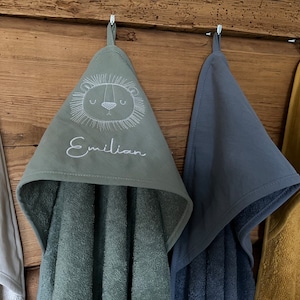 May include: Four hooded towels hanging on hooks. The towel in the center has a lion face and the name "Emilian" embroidered on it. The towels are in white, green, blue, and yellow.