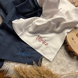 May include: Two personalized towels, one navy blue with the name "Leo" embroidered in blue thread, and one white with the name "Ulrike" embroidered in red thread.