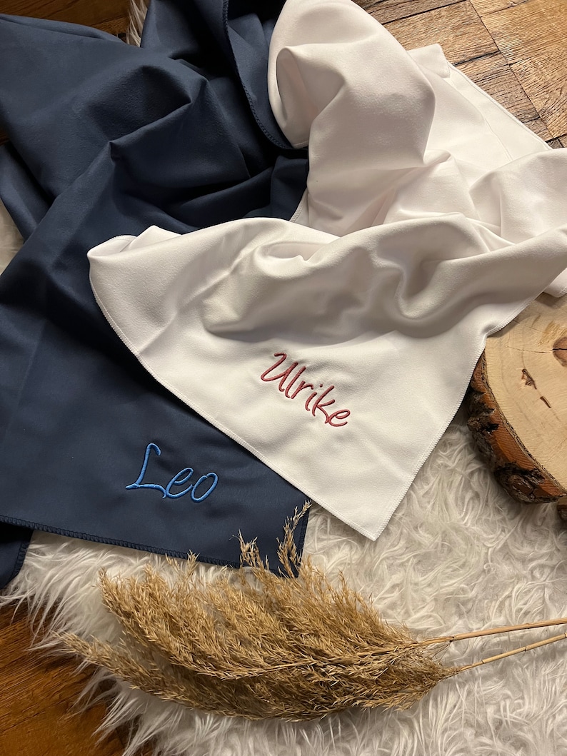 May include: Two personalized towels, one navy blue with the name "Leo" embroidered in blue thread, and one white with the name "Ulrike" embroidered in red thread.