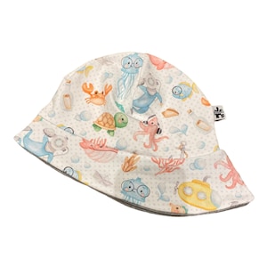 Op de afbeelding: Een witte zonnehoed met een grijze rand en een kleurrijke print met een onderwaterthema. De print bevat zeedieren zoals een schildpad, octopus, kwal en een krab. Er zijn ook kleine witte stippen op de hoed.