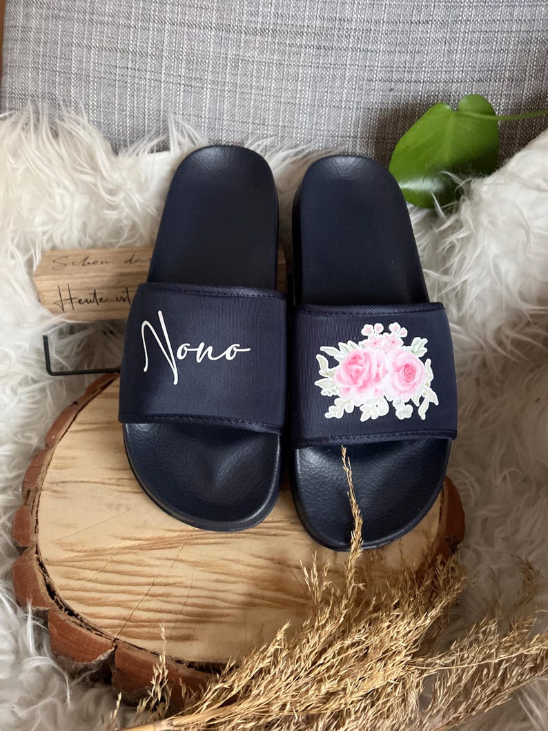 May include: A pair of navy blue slides with a floral embroidery design. One slide has the word "Jono" embroidered on the strap.