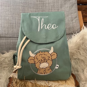 Könnte beinhalten: Ein grüner Rucksack mit Kordelzug und einem niedlichen gestickten Hochlandrind-Motiv sowie dem Namen "Theo", der auf der Vorderseite gestickt ist.