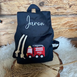 Può includere: Uno zaino in tela nera con un nome ricamato in bianco "Jaron" e un camion dei pompieri ricamato in rosso con il numero "112" sul davanti.