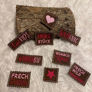 Könnte beinhalten: Neun braune Filzpatches mit rosa gesticktem Text und Designs. Die Patches beinhalten die Wörter "Cool Kids", "Herzstück", "Wildfang", "Wunderbar", "Wunsch Kind", "Frech Dachs", "Frech Wild Wunderbar" sowie ein Herz und einen Stern.