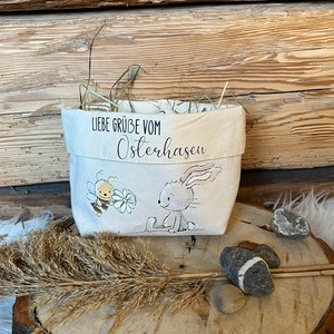K&ouml;nnte beinhalten: Eine wei&szlig;e Stofftasche mit einem Hasen- und Bienen-Motiv. Die Tasche hat den Text "Liebe Gr&uuml;&szlig;e vom Osterhasen" aufgedruckt.