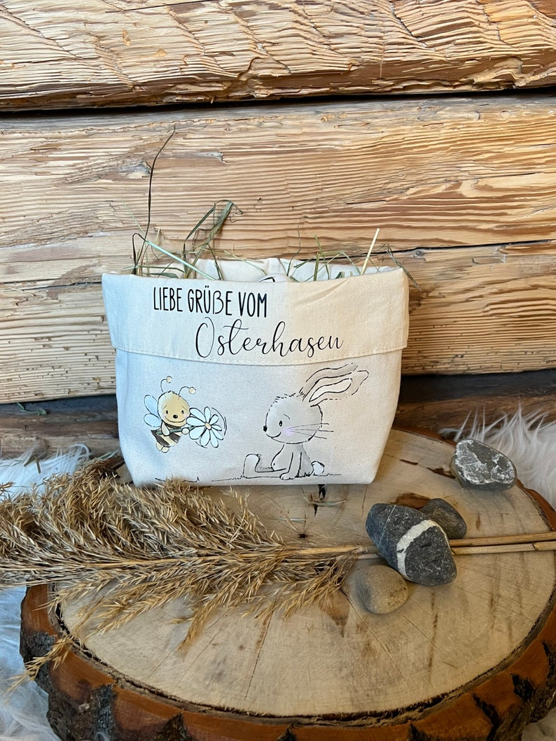 K&ouml;nnte beinhalten: Eine wei&szlig;e Stofftasche mit einem Hasen- und Bienen-Motiv. Die Tasche hat den Text "Liebe Gr&uuml;&szlig;e vom Osterhasen" aufgedruckt.