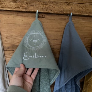 May include: Three hooded towels hanging on hooks. The towel in the center is green with a white embroidered lion face and the name "Emilian". The towel on the left is white and the towel on the right is blue.