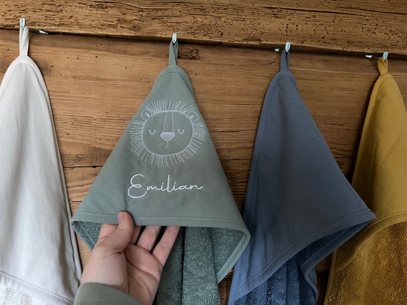 May include: Three hooded towels hanging on hooks. The towel in the center is green with a white embroidered lion face and the name "Emilian". The towel on the left is white and the towel on the right is blue.