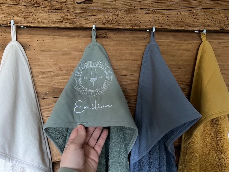 May include: Four hooded towels hanging on hooks. One towel is white, one is green with a lion face and the name "Emilian" embroidered on it, one is blue, and one is yellow.