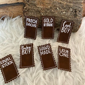 Può includere: Nove toppe di tessuto marrone con testo ricamato bianco. Il testo sulle toppe recita: "Frech Dachs", "Gold Stück", "Cool Boy", "Baby Boy", "Hand Made", "Little One", "Einzel Stück".