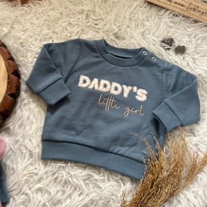 Könnte beinhalten: Blaues Baby-Sweatshirt mit dem Text "DADDY'S little girl" in weißer und goldener Schrift. Das langärmlige Oberteil hat einen Rundhalsausschnitt und Druckknöpfe an der Schulter. Das Kleidungsstück wird auf einem weißen, flauschigen Teppich präsentiert.