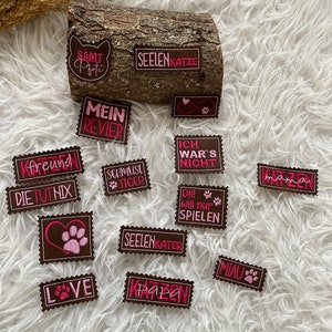 Peut inclure: Un ensemble de patchs en feutre marron avec des broderies roses. Les patchs présentent diverses phrases sur le thème du chat en allemand, notamment "Seelenkatze", "Mein Revier", "Die tut nix", "Schmuse Tiger", "Seelenkater", "Ich war's nicht", "Die will nur spielen", "Mama", "Freund", "Papa", "Mia", et "Love".