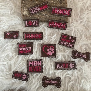 Può includere: Una collezione di toppe in feltro marrone con testo ricamato rosa. Le toppe presentano frasi come "Seelenhund", "Freund", "Love", "Mama", "Hunde", "Ich war's nicht", "Wuff", "Mein Revier", "Der will nur spielen", "Dertutnix", "Leckerli" e "Impfpass".
