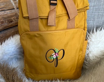 Könnte beinhalten: Ein gelber Canvas-Rucksack mit braunen Trägern und einer schwarzen Schnalle. Der Rucksack hat einen Klappverschluss und eine Reißverschlusstasche auf der Vorderseite. Die Vorderseite des Rucksacks ist mit einem Blumendesign und dem Buchstaben "P" bestickt.