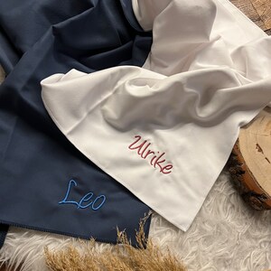 May include: Two personalized towels, one navy blue with the name "Leo" embroidered in blue thread, and one white with the name "Ulrike" embroidered in red thread.
