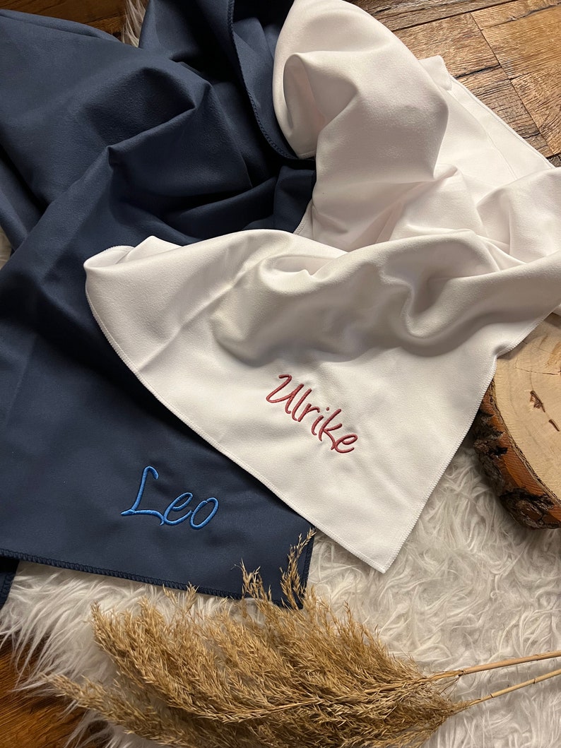 May include: Two personalized towels, one navy blue with the name "Leo" embroidered in blue thread, and one white with the name "Ulrike" embroidered in red thread.